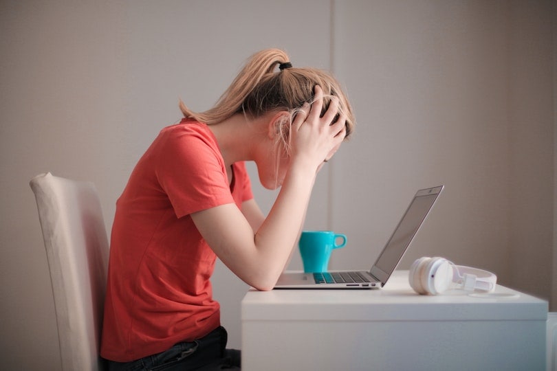 woman in red shirt holding her head while looking at her laptop