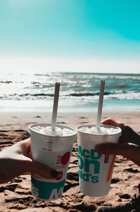 two persons holding cups of mcdonald's drinks at the beach