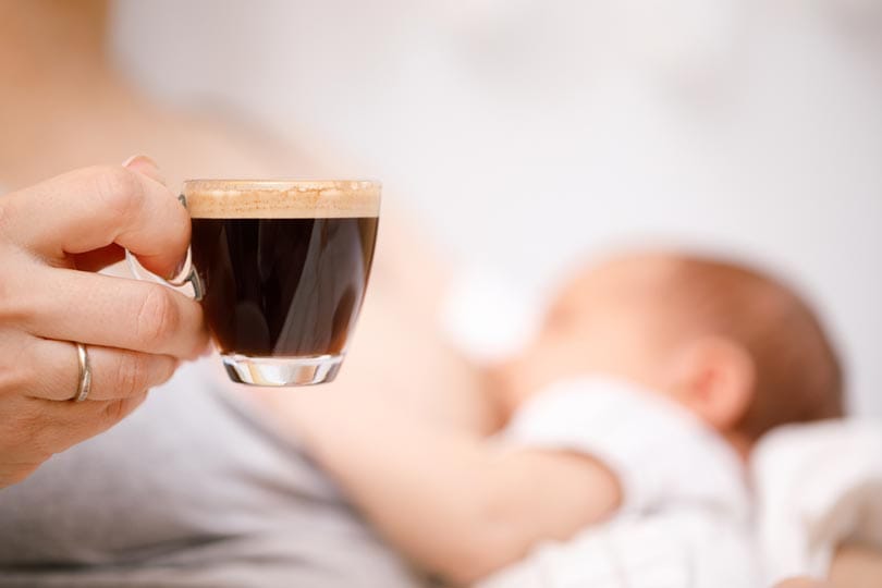 mother breastfeeding newborn baby while drinking coffee