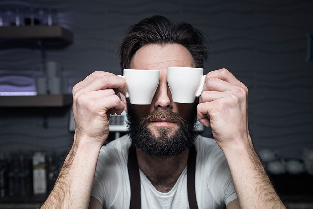 bearded man covering his eyes with cups of coffee